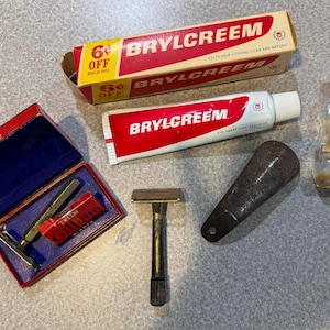 Puede incluir: Un surtido de productos de aseo personal vintage, que incluye un tubo de Brylcreem, una maquinilla de afeitar Gillette en una caja roja, una brocha de afeitar y una botella de champú Wildroot Liquid Cream. Los artículos están dispuestos sobre una superficie gris.