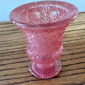 May include: A small, translucent pink glass vase featuring a pressed pattern of geometric stars and diamonds. The vessel has a wide, flared rim, a tapered body, and a circular pedestal base with a radiating ribbed design on the bottom edge.