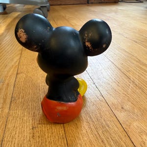May include: A vintage Mickey Mouse figurine, viewed from the back, with black ears and head, a black torso, red shorts, and yellow gloves. The figurine shows signs of wear and tear, with paint loss on the ears.