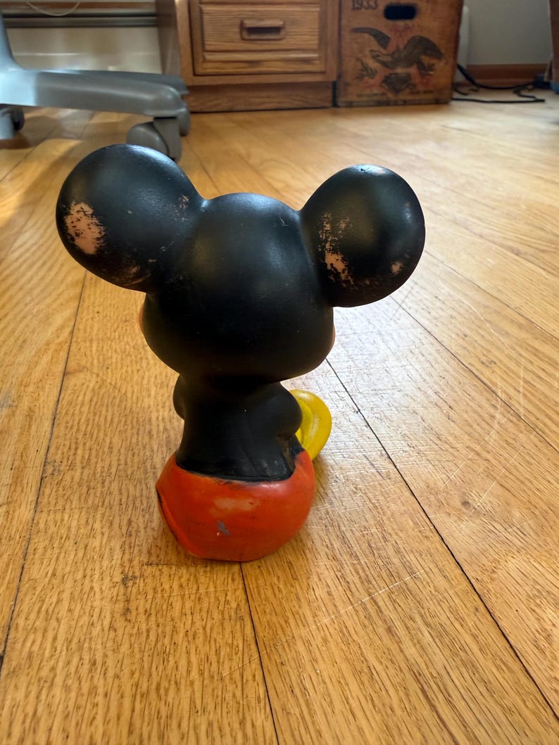 May include: A vintage Mickey Mouse figurine, viewed from the back, with black ears and head, a black torso, red shorts, and yellow gloves. The figurine shows signs of wear and tear, with paint loss on the ears.