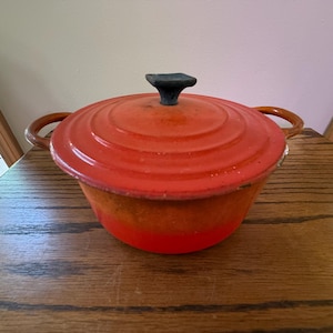 May include: A round, orange enameled cast iron Dutch oven with a matching lid. The pot features two side handles and a dark, square-shaped knob on the center of the lid. The exterior displays a gradient of orange tones, transitioning from a lighter shade at the top to a vibrant, saturated orange at the base.