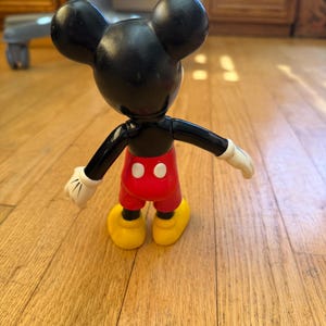 Op de afbeelding: Een vintage Mickey Mouse-figuur, van achteren gezien, met zwarte oren en hoofd, rode broek, gele schoenen en witte handschoenen. De figuur staat op een houten vloer.
