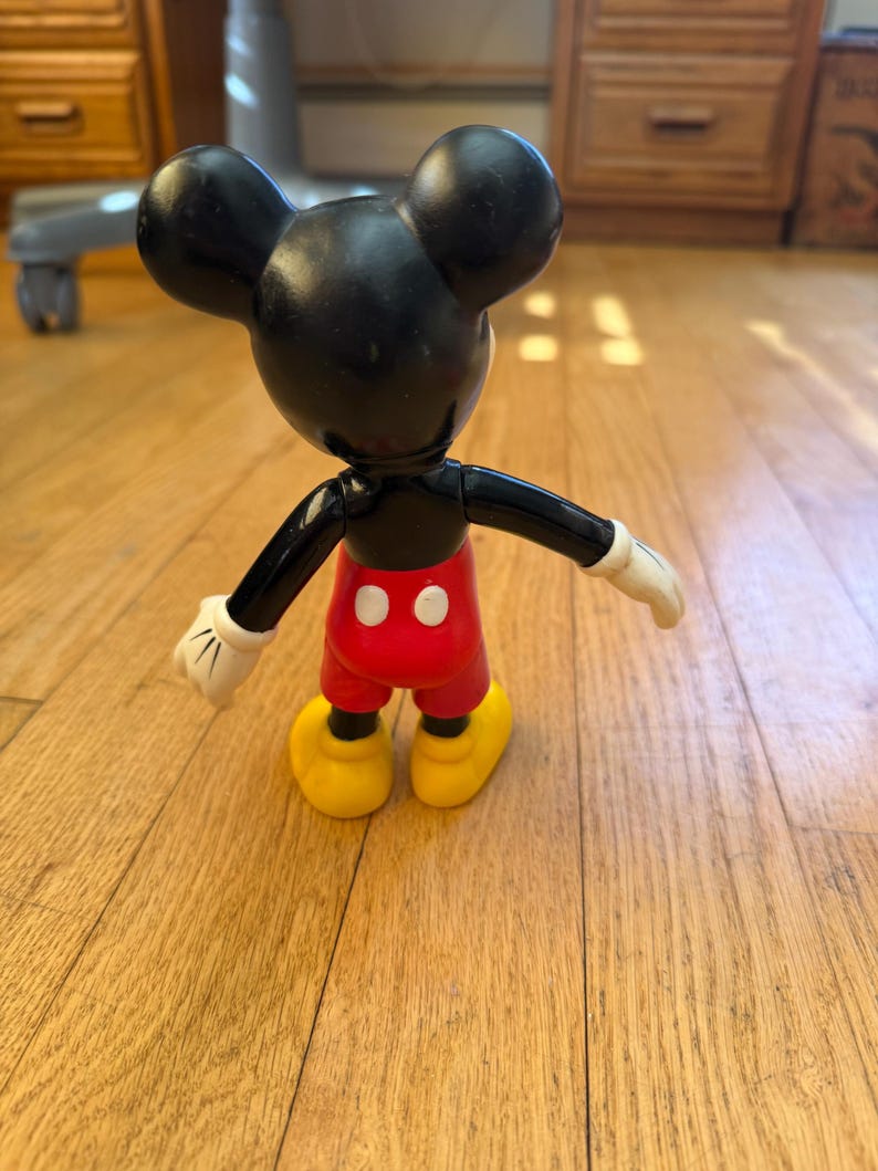 Op de afbeelding: Een vintage Mickey Mouse-figuur, van achteren gezien, met zwarte oren en hoofd, rode broek, gele schoenen en witte handschoenen. De figuur staat op een houten vloer.
