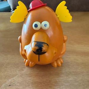 Könnte beinhalten: Orangefarbener Spielzeughund aus Kunststoff mit rotem Hut und gelben Ohren. Das Spielzeug hat blaue Augen und eine schwarze Nase. Der Hund sitzt auf einer Holzoberfläche.