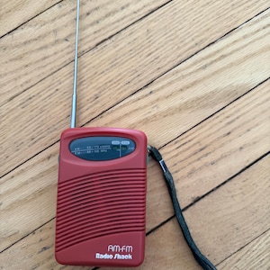 May include: A red AM-FM Radio Shack portable radio with a retractable antenna and a black wrist strap. The radio has a speaker grill design and a display showing the frequency range. The Radio Shack logo is visible.