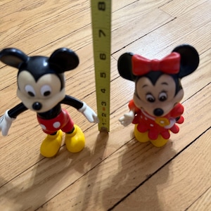 Op de afbeelding: Twee Disney-beeldjes van Mickey Mouse en Minnie Mouse worden getoond. Mickey draagt een rode broek, gele schoenen en witte handschoenen. Minnie draagt een rode jurk met een rode strik. De figuren zijn ongeveer 7.6 cm hoog.