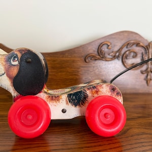 Op de afbeelding: Vintage houten trekdier speelgoed in de vorm van een hond met een geschilderde gevlekte vacht, zwarte oren en rode wielen. De hond heeft een groene knopstaart en een zwart trekkoord.
