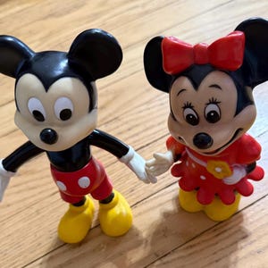 Op de afbeelding: Twee vintage Mickey Mouse en Minnie Mouse beeldjes. Mickey draagt een klassieke outfit met een rode broek, gele schoenen en witte handschoenen. Minnie draagt een rode jurk met witte stippen en een rode strik.