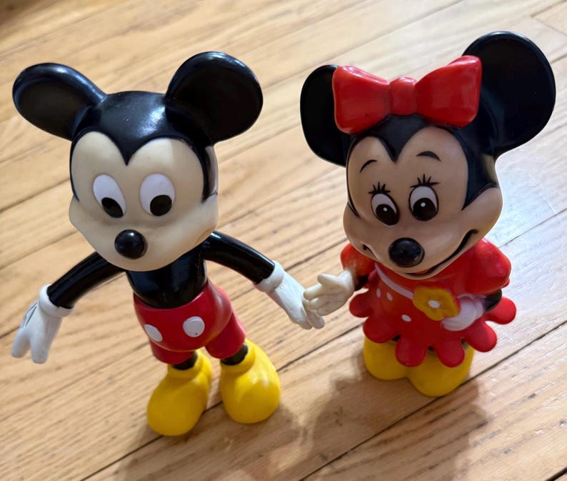 Op de afbeelding: Twee vintage Mickey Mouse en Minnie Mouse beeldjes. Mickey draagt een klassieke outfit met een rode broek, gele schoenen en witte handschoenen. Minnie draagt een rode jurk met witte stippen en een rode strik.