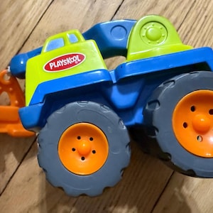 May include: A colorful Playskool toy truck with a blue chassis, gray wheels with orange centers, and a yellow and green cab. The truck has an orange plow and the Playskool logo.