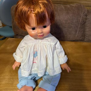 May include: A vintage doll with auburn hair styled in a bun. The doll wears a white blouse with a floral design and light blue pants. The doll is sitting on a wooden surface.
