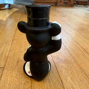 May include: A black figurine of a mouse wearing a top hat and tuxedo. The mouse is posed with one hand raised to its hat. The figurine has white gloves and a red button on its tail.