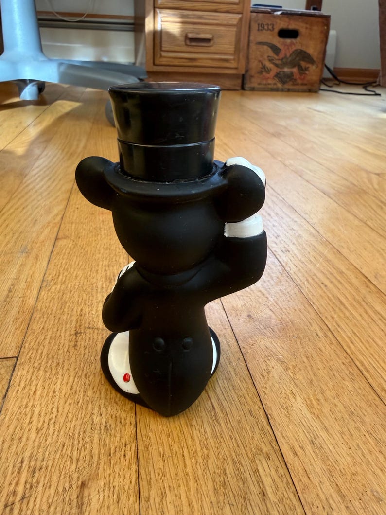 May include: A black figurine of a mouse wearing a top hat and tuxedo. The mouse is posed with one hand raised to its hat. The figurine has white gloves and a red button on its tail.