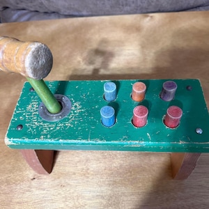 May include: A vintage wooden toy with a green base, featuring six colorful pegs and a wooden mallet. The mallet has a light brown head and a green handle. The base is supported by two wooden legs.