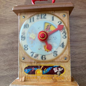 May include: Vintage Fisher-Price toy clock with a red handle and clock hands. The clock face features numbers, musical notes, and a cartoon mouse. The base of the clock has the words "Fisher Price Toys".