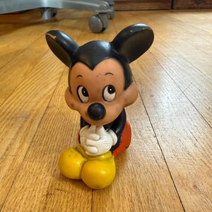 May include: A vintage Mickey Mouse figurine. The figure has a black head and ears, a tan face, a black nose, and white gloves. It is wearing a red shorts and yellow shoes. The figurine is sitting on a wooden floor.