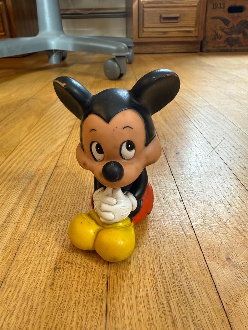 May include: A vintage Mickey Mouse figurine. The figure has a black head and ears, a tan face, a black nose, and white gloves. It is wearing a red shorts and yellow shoes. The figurine is sitting on a wooden floor.