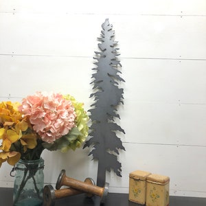May include: A dark gray metal tree silhouette stands tall, next to a bouquet of pink and yellow flowers in a glass jar. Two wooden spools and two small, yellow floral-decorated tins complete the arrangement. The items are arranged on a dark surface against a white wood-paneled wall.