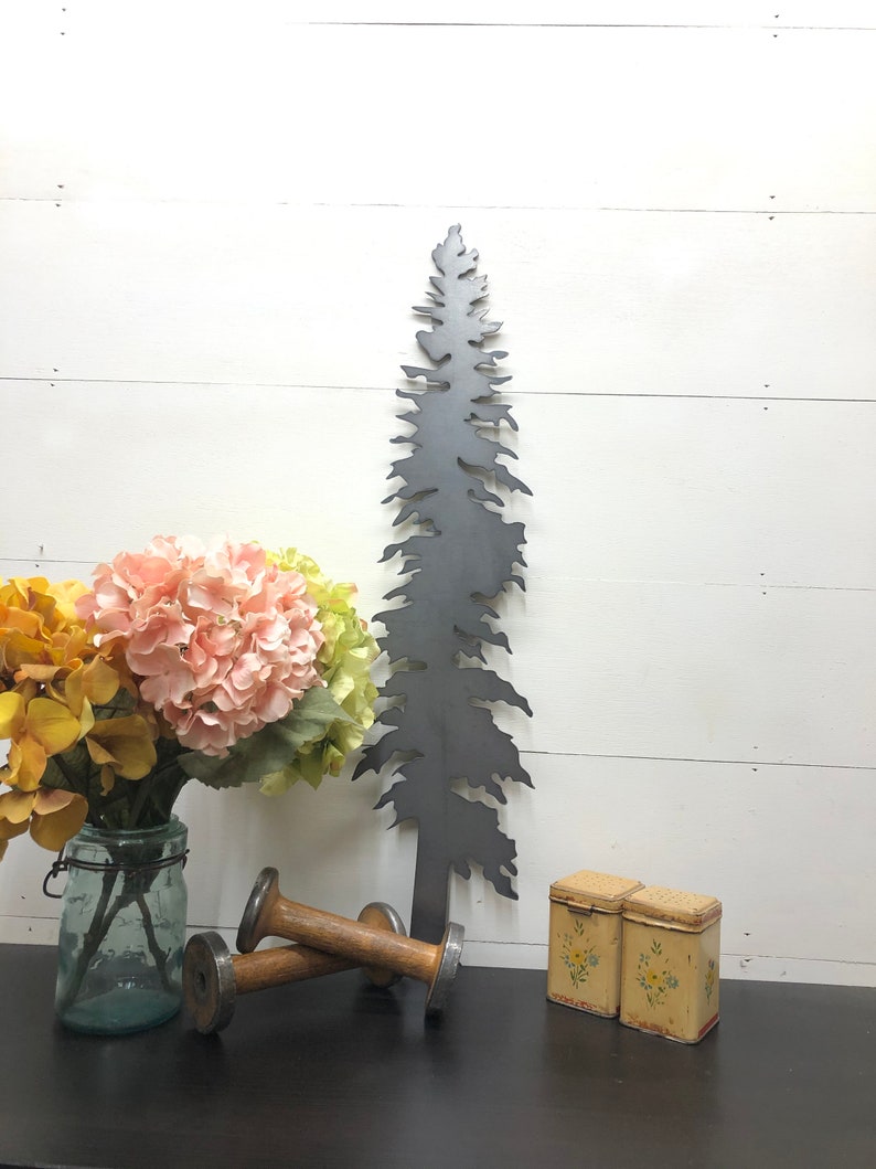 May include: A dark gray metal tree silhouette stands tall, next to a bouquet of pink and yellow flowers in a glass jar. Two wooden spools and two small, yellow floral-decorated tins complete the arrangement. The items are arranged on a dark surface against a white wood-paneled wall.