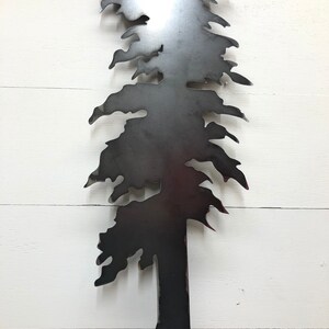 May include: A metal tree silhouette with a dark gray finish. The tree has a detailed design with branches and needles, and is mounted against a white wooden background. The tree's trunk is visible at the bottom, and the overall design is simple and elegant.