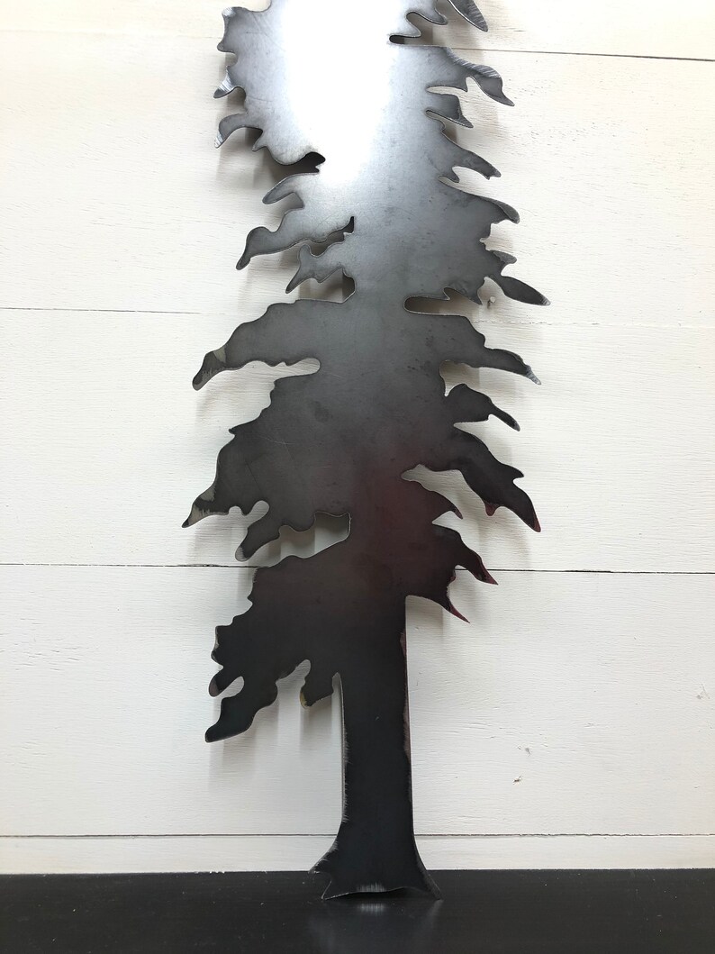 May include: A metal tree silhouette with a dark gray finish. The tree has a detailed design with branches and needles, and is mounted against a white wooden background. The tree's trunk is visible at the bottom, and the overall design is simple and elegant.