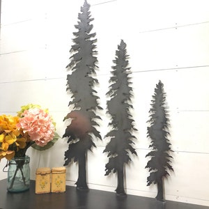 May include: Three metal tree silhouettes of varying heights are displayed against a white wooden background. The trees are cut from dark metal and have detailed, textured edges. The scene includes a black surface with a vase of flowers and two small decorative boxes.