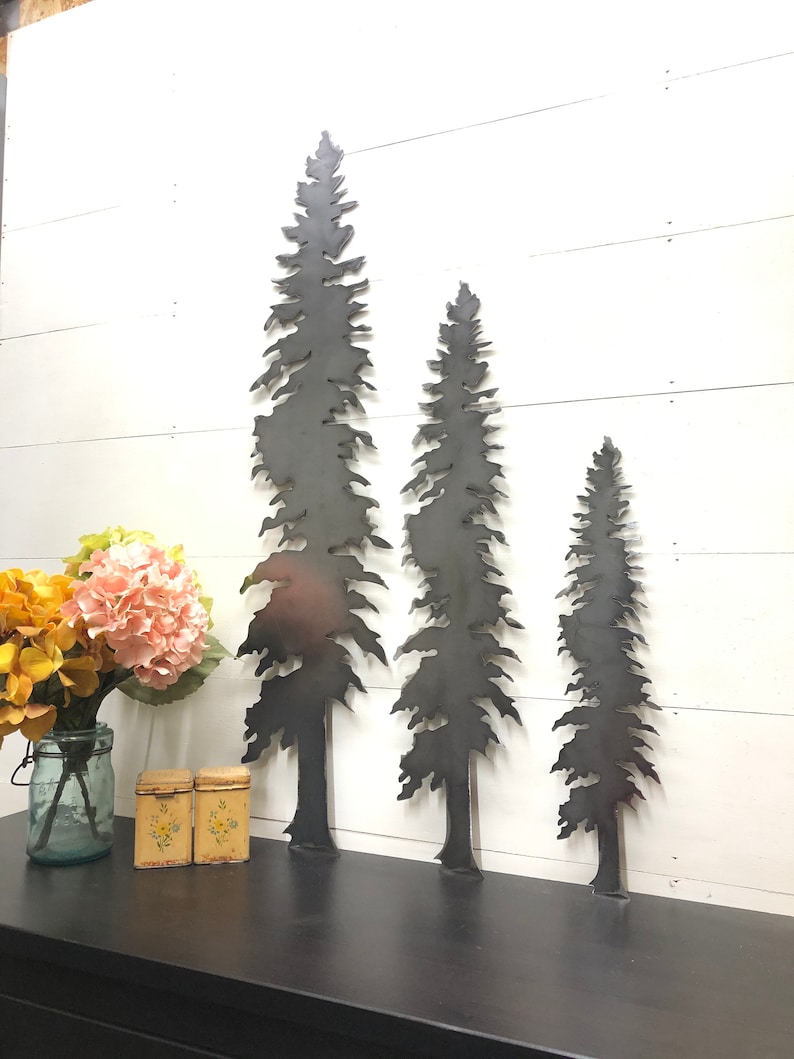 May include: Three metal tree silhouettes of varying heights are displayed against a white wooden background. The trees are cut from dark metal and have detailed, textured edges. The scene includes a black surface with a vase of flowers and two small decorative boxes.