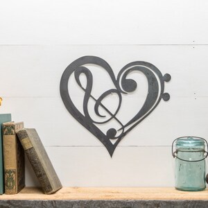 May include: A metal heart-shaped wall decor with a treble clef and bass clef design. The decor is painted in a dark gray color and is displayed on a white wall.