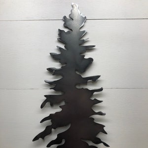 May include: A dark gray metal tree silhouette, cut from a single sheet of metal. The tree has a detailed, layered design with a textured appearance. The artwork is mounted on a white wooden background, creating a contrast.