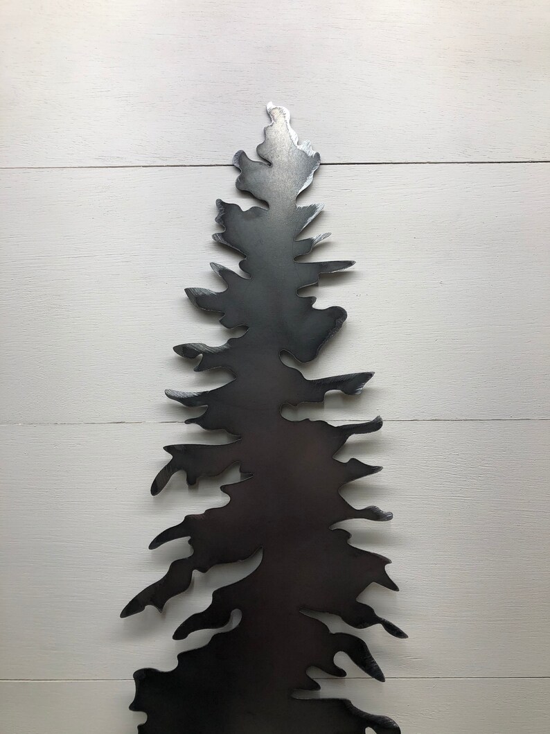 May include: A dark gray metal tree silhouette, cut from a single sheet of metal. The tree has a detailed, layered design with a textured appearance. The artwork is mounted on a white wooden background, creating a contrast.