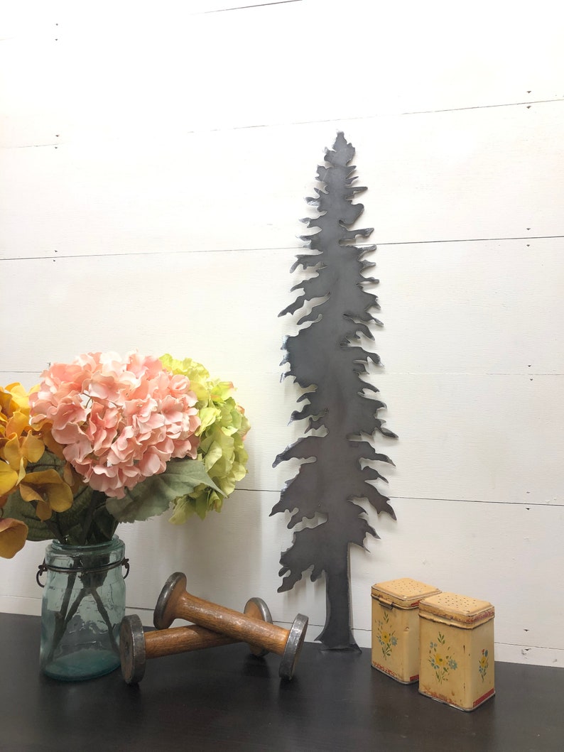 May include: A dark metal tree silhouette stands tall against a white wood-paneled wall. A bouquet of pink and yellow flowers in a glass jar, two wooden spools, and two small, floral-decorated boxes complete the rustic decor.