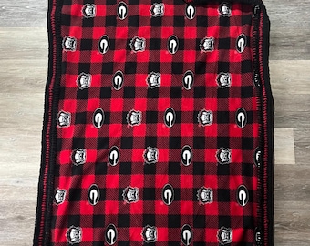 May include: Red and black plaid blanket with a black crocheted border. The blanket features a repeating pattern of black and white emblems. The blanket is made of a soft material and is folded on a wooden surface.
