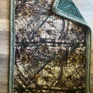 May include: A rectangular camouflage-patterned blanket with a green crocheted border. The blanket features a forest design in shades of brown, green, and tan. The reverse side is a solid dark green, textured fabric. The blanket is designed for comfort and warmth.