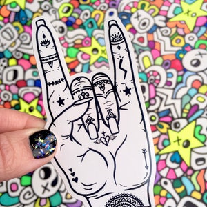 May include: A white sticker with a black line drawing of a hand giving the rock and roll hand gesture. The hand has several tattoos including a heart with wings, a star, a lightning bolt, and a mandala.