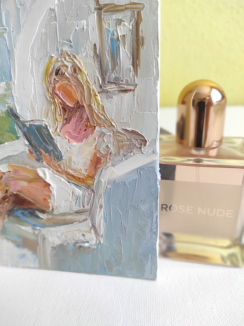 May include: A painting of a woman reading a book in a white room with a window. The painting is done in a loose, impressionistic style with thick brushstrokes. The woman is wearing a white shirt and has blonde hair. The painting is framed and has a light blue background.