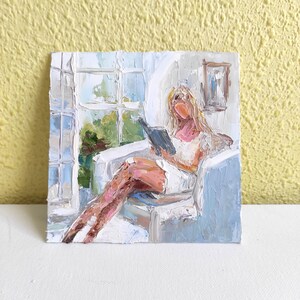 May include: A painting of a person sitting in a chair by a window, reading a book. The person is wearing a white dress and has blonde hair. The painting is done in a loose, impressionistic style.