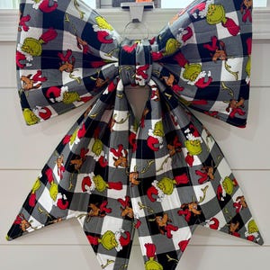 May include: A large fabric bow featuring a black and white buffalo plaid pattern with a repeating print of a green character in a red Santa hat and a brown reindeer. The bow has two wide top loops and two long, pointed tails hanging downwards.