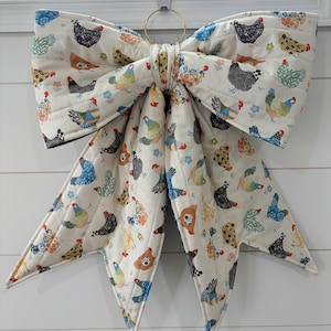 May include: A large, quilted fabric bow featuring a repeating pattern of colorful chickens on an off-white background. The bow is constructed with two wide top loops and two long, pointed tails, all held together by a central fabric knot and a gold ring.