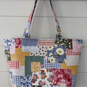 May include: A quilted tote bag featuring a patchwork design of various fabric squares. The patterns include floral prints in red, blue, and yellow, red and white gingham, blue and white checks, and striped sections, all stitched together.