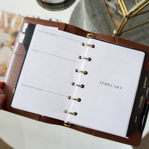 May include: A brown leather bound planner with gold rings. The planner is open to the February page. The page has a section for "Priorities & Goals", "To Do", and "Improve Next Month". The right side of the planner has tabs for "Finance", "Personal", and "Misc."