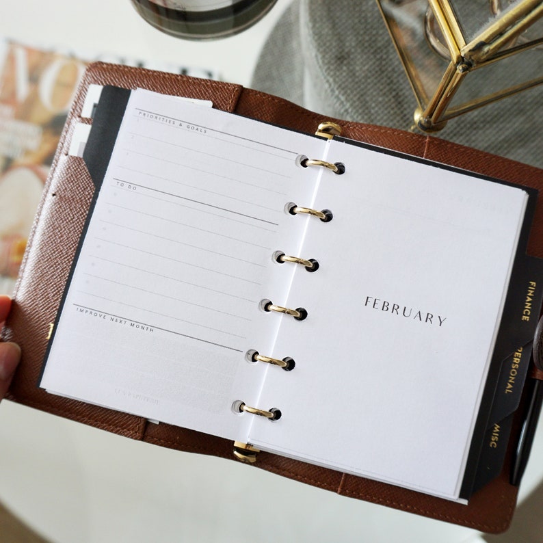May include: A brown leather bound planner with gold rings. The planner is open to the February page. The page has a section for "Priorities & Goals", "To Do", and "Improve Next Month". The right side of the planner has tabs for "Finance", "Personal", and "Misc."