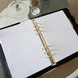 May include: Open weekly planner with gold-colored rings and a black leather cover. The planner is open to a weekly spread with sections for each day of the week. A glass of wine and a jar of gold paper clips are in the background.