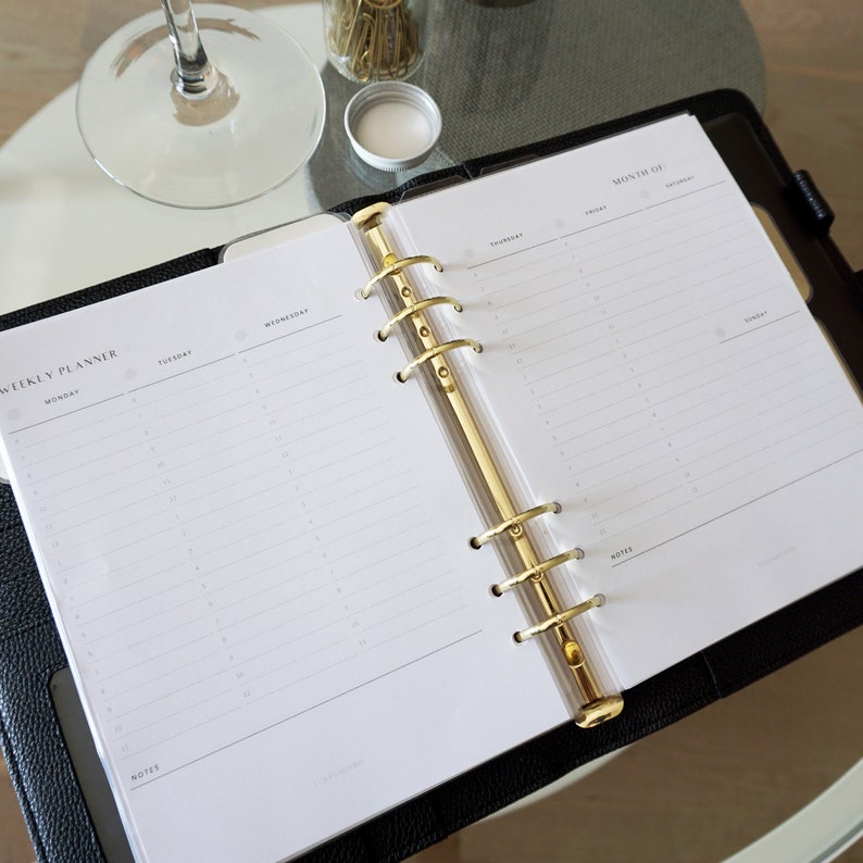 May include: Open weekly planner with gold-colored rings and a black leather cover. The planner is open to a weekly spread with sections for each day of the week. A glass of wine and a jar of gold paper clips are in the background.