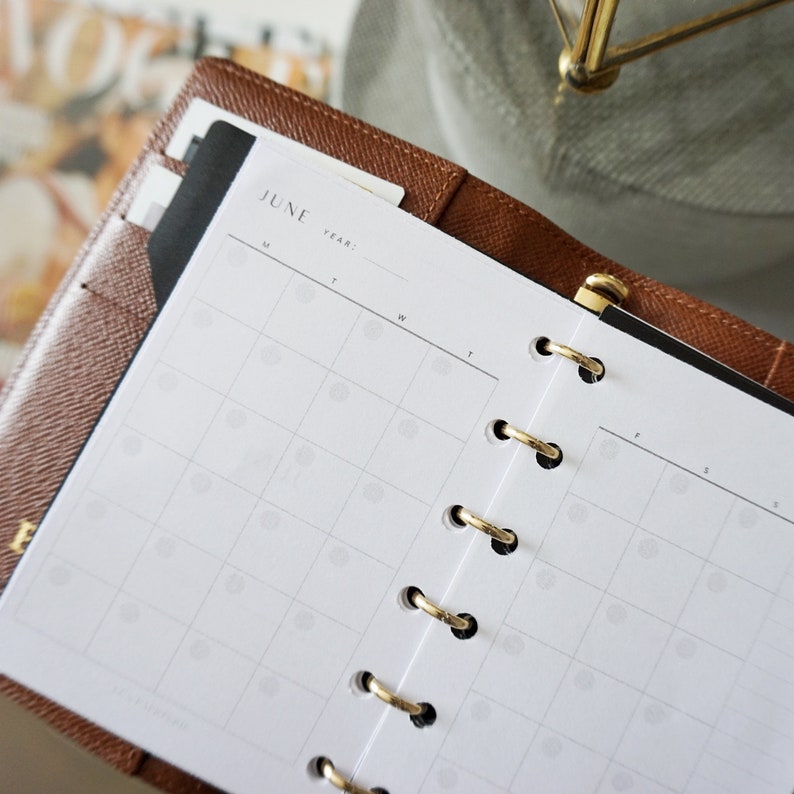 May include: A brown leather bound planner with gold rings and a white monthly calendar insert for June. The calendar has a grid layout with spaces for each day of the month.