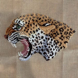 May include: A needlepoint embroidery of a leopard head in profile, featuring a tan coat with black spots, a green eye, and an open mouth with white teeth and pink gums. The design includes white whiskers and is stitched onto a tan mesh fabric.