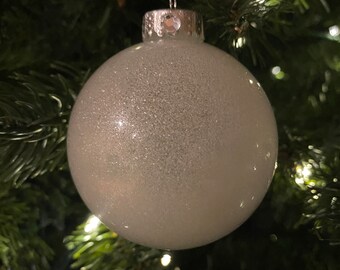 May include: A spherical, silver Christmas bauble with a sparkly surface hangs from a white ribbon. The decoration is set against a backdrop of green Christmas tree branches and fairy lights.