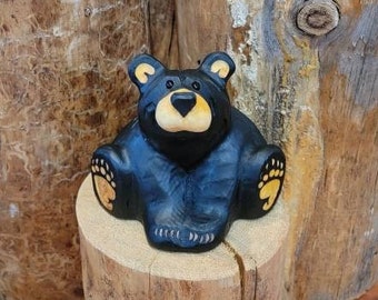 May include: A black bear wood carving with yellow painted paws sits on a wooden log. The bear is sitting up with its paws in front of it. The carving is on a wooden log with a rough texture.