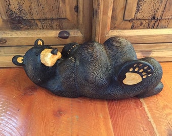 May include: A carved wooden black bear sculpture lying on its back with its paws in the air. The bear is painted black with a brown nose and a light brown paw print on its back.