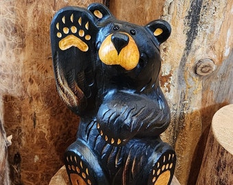 May include: A black wooden carved bear figurine with a raised paw, sitting on a wooden stump. The bear has a friendly expression and is painted with yellow accents on its paws and nose.