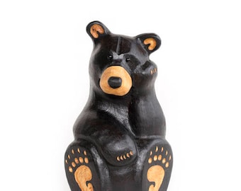 May include: A black wooden bear figurine with a brown nose and paw prints. The bear is sitting with its paws tucked in front of it.